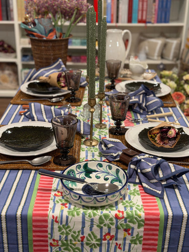 Blue and white tablescape with geranium tablerunner, Spanish grating plate, white dishes with green leaf appetizer plates, purple wine glasses, and green candles in brass candle holder