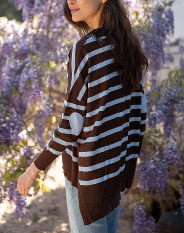 A woman wearing a brown and white horizontally striped knit poncho sweater with a white elbow patch, standing outdoors in front of purple wisteria flowers.