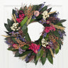 A round wreath made of dried flowers including shades of pink, purple, green, and beige, arranged in a wooden crate against a white wooden background.