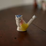 A small ceramic bird-shaped pipe or ashtray with a white body, yellow belly, pink chest, and blue hat, sitting on a dark wooden tabletop.
