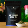 August Olympus Mint Cucumber Herbal Tea package with tea leaves in a bowl on a wooden surface.