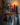This image shows a shelf with hooks upon which sits a Greentree 8” Petite Pillar candle, lit, shown in red. It sits amongst wintery items.