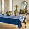 A set dining table with a blue tablecloth featuring a hydrangea pattern, surrounded by window light and decorative room elements.