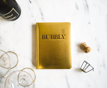 Gold book titled 'Bubbly' on a marble surface with champagne bottle and glasses.