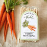 Packaged carrot fettuccine with a Valente Market label next to fresh carrots on a textured surface.