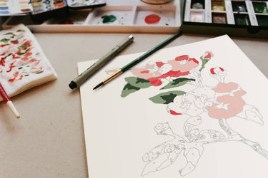 Artistic drawing of a character with flowers on paper, with watercolor paints and brushes in the background.
