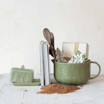 Image of holiday themed kitchen items, green debossed tree butter dish, green debossed tree batter bowl in which sits two small wrapped presents, a tinsel tree and two wooden spoons. Two books are propped between the butter dish and bowl. A small pile of ground cinnamon is in the foreground.