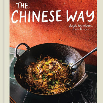 Book cover features a terracotta orange wall and a dark green tile surface on which sits a carbon steel wok filled with a mushroom and noodle stir fry. There is wok tool in the wok and a pair of chopsticks lies in the foreground on the counter.