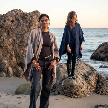 Two models standing on a rocky beach. The model in the foreground is wearing the hazelnut with navy windowpane plaid, the wrap is open. The model in the background is standing on a rock and she has the Navy with red windowpane plaid wrapped around her body.