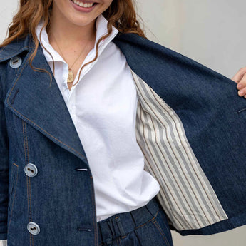 Smiling model is holding open one side of the denim Peacoat to reveal the striped inner lining. She is wearing a white blouse with the collar popped under the peacoat.