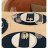 A styled dining table setting featuring navy blue oval placemats, white plates, a navy napkin with silver cutlery, and clear drinking glasses, with a brown leather upholstered chair in the background.