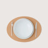A minimalist tabletop setting featuring a light brown oval placemat with a white ceramic dinner plate and matching white cutlery, photographed from directly above on a light beige background.