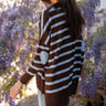 A woman wearing a brown and white horizontally striped knit poncho sweater with a white elbow patch, standing outdoors in front of purple wisteria flowers.