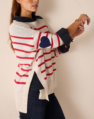 A woman wearing a cream knit sweater with red horizontal stripes, dark navy cuffs, and a navy heart patch on the sleeve, paired with dark jeans against a plain beige wall.