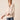 A woman modeling a light beige long-sleeve top with a split V-neckline and buttoned cuffs, paired with blue jeans, against a solid light beige background.